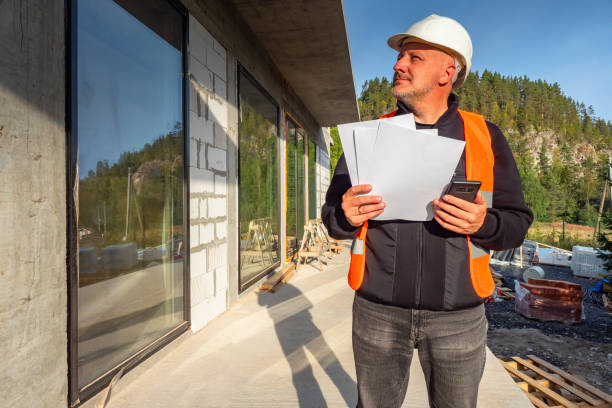 istock Architect at a construction site. Man in a construction worker uniform holds a telephone. Architect near a building under construction. Concept - architect wants to call a customer. Panoramic windows 2159800061