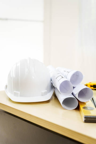 An architect's desk with blueprints and a white hardhat sits empty, ready for the next project. No people are present, highlighting the tools of the trade.