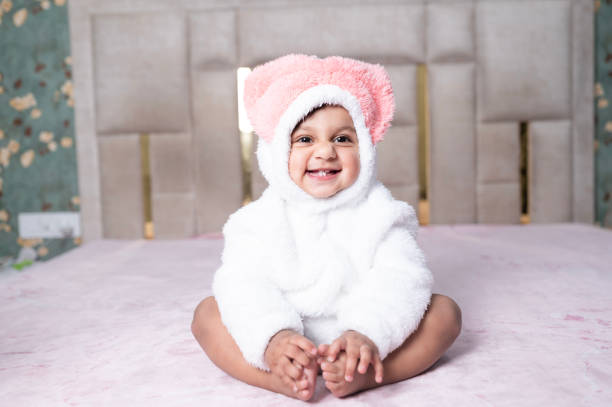 A charming baby dressed in a fluffy white costume with a large pink bow, sitting on a soft pink surface. The baby is smiling brightly, showcasing a joyful and heartwarming moment.