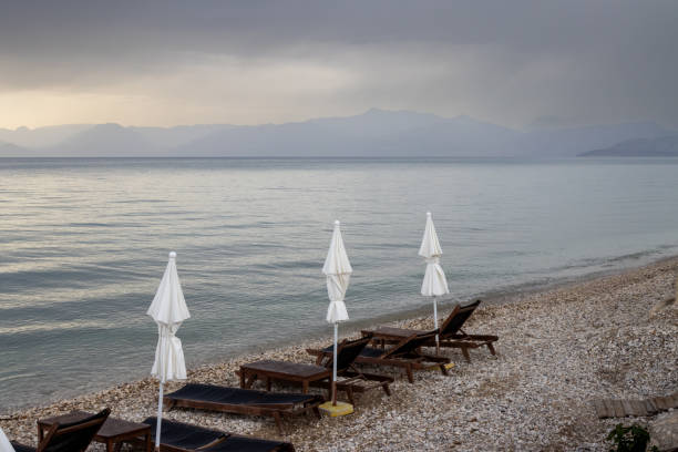 Beach chairs and umbrellas in the evening. Calm sea during a sunset in the late spring. Cloudy sky. Coast of Albania with high mountains on the horizon. Acharavi, Korfu, Greece.