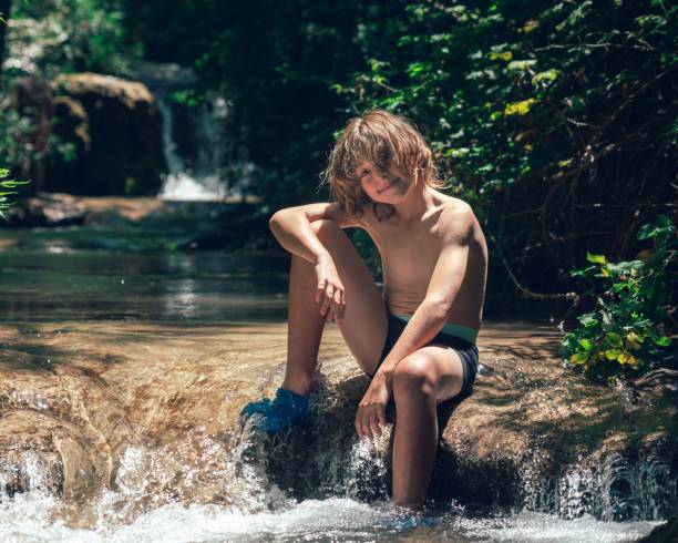 istock Young boy sitting on a rock in a serene forest stream on a sunny day 2159828765