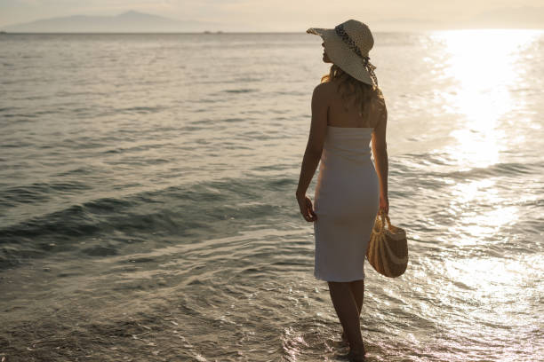 Rear view shot of young  woman in white dress and straw hat standing on the beach and enjoying sunset above the sea