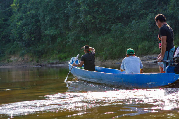 Kwamalasamutu, Suriname – November 24, 2016: Three indigenous men are sailing in a canoe with an outboard motor on a river in the tropical rainforest. The boatman shields his eyes from the sun.