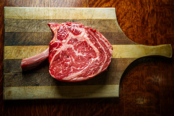 Raw Tomahawk Rib Eye Beef Steak on a Rustic Wooden Cutting Board Ready to Cook Next to a Charcoal Grill in a Residential Back Yard in Summertime