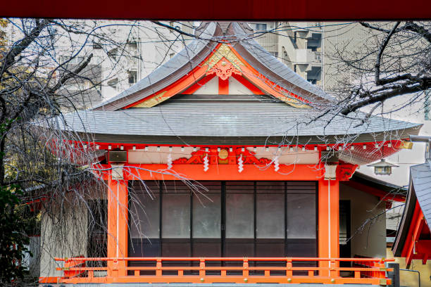 Frontal view of the Hanazono Shrine, a 17th Shinto shrine, with mostly bare tree branches in the front and tall building showing in the background, located in Shinjuku, Tokyo, Japan