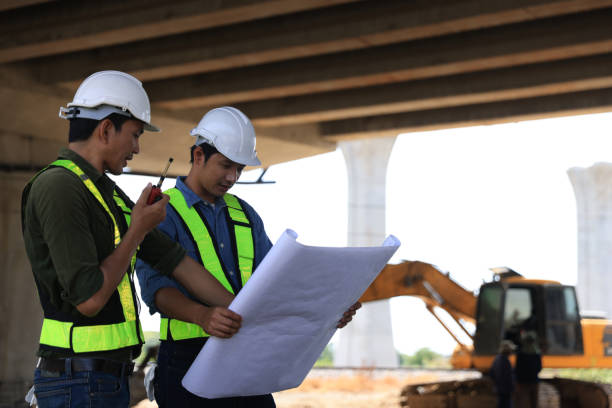 istock Two male  construction engineers  in safety vests and helmets discussing plans under the motorway  bridge at the construction site 2159842965