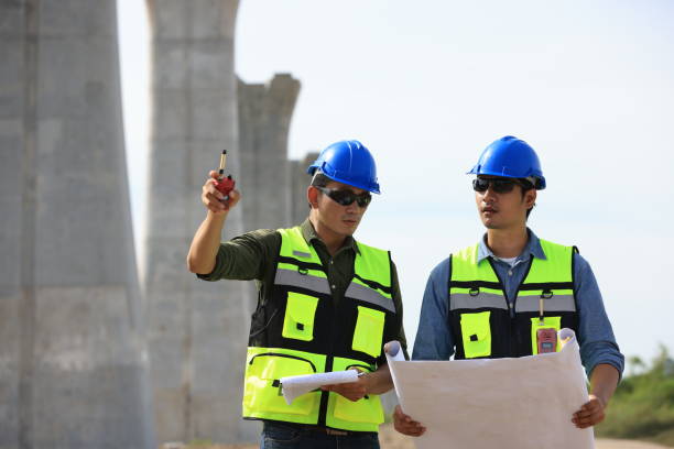 istock Survey construction technicians wearing safety vests and hard hats work with level survey cameras at road construction  site 2159842970