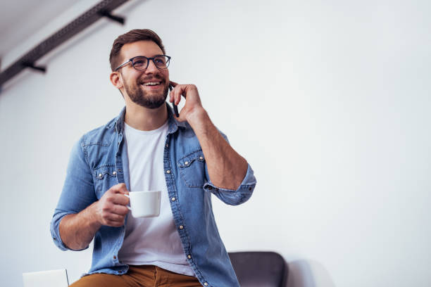istock Handsome bearded man leaning on office desk, drinking coffee and talking on the phone. 2159849985