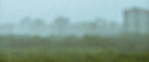 Atmospheric minimal backdrop with rain droplets on glass. Wet window with rainy drops and dirt spots closeup. Blurry buildings and green trees under blue sky against dirty window glass with raindrops.