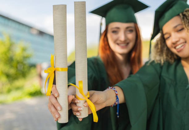 Two young women in green graduation gowns, proudly holding their diplomas with yellow ribbons, celebrating their achievement.