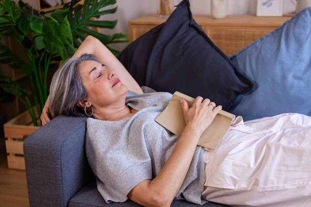 istock Senior woman relaxing on sofa holding book falling asleep 2159901045