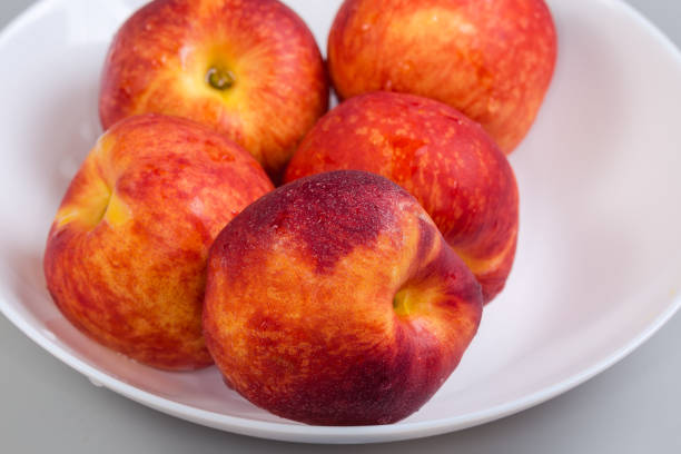 Ripe nectarines covered with dew drops on the white dish on a gray background, fragment close-up in selective focus