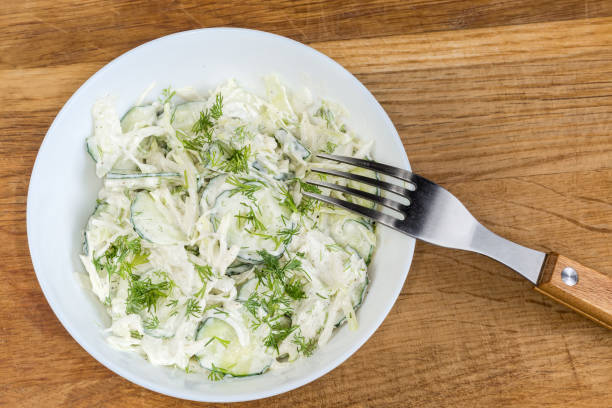 Green vegetable salad made of sliced fresh young cabbage, cucumbers, onion, dill with mayonnaise dressing in the white bowl with fork on an old oak wooden surface, top view