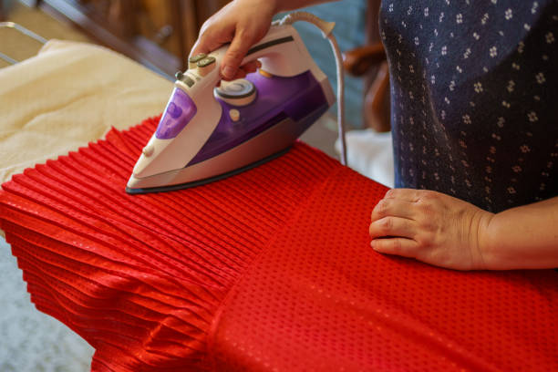 Close-up of a hand using a steam iron on a red textured fabric on an ironing board. Concept of household chores and fabric care.