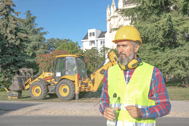 A construction professional in an orange vest and hard hat points towards heavy machinery, overseeing its operation in a bustling construction environment.