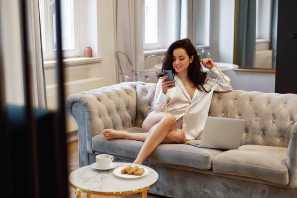 istock Smiling young lady using smartphone while sitting on the sofa in living room while working on laptop online. Technology, domestic lifestyle concept 2159925872