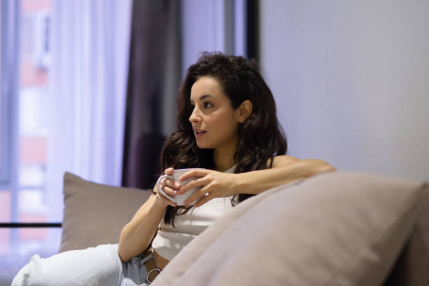 Close up of beautiful young woman sitting on sofa indoor at modern home looking away watching TV holding in hand cup of coffee. Attractive female resting in appartment with hot drink in hand. primer plano de una mujer joven bonita sentada en el sofá interior de una casa moderna que mira hacia otro lado viendo la televisión sosteniendo en la mano una taza de café. hembra atractiva descansando en apartamento con bebida caliente