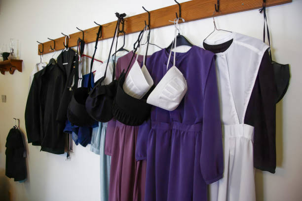 Amish clothing hanging on wooden rack: plain dresses, coats, and bonnets. Amish clothing on wooden rack: plain, modest garments. Purple dresses, black coats, white bonnets. Represents Amish culture, traditional religious attire, simple living, and faith-based dress codes. Amish stock pictures, royalty-free photos & images