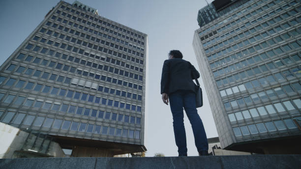 A Caucasian senior adult in business attire carries a briefcase while walking near modern office skyscrapers, reflecting a busy urban work environment.