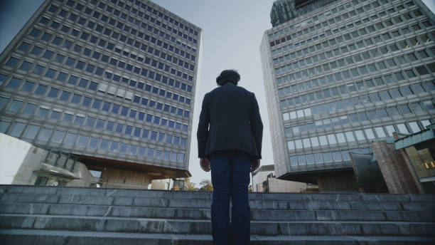 Elderly Caucasian man in formal attire strides confidently across a plaza, with towering office buildings lining his path under a clear sky.