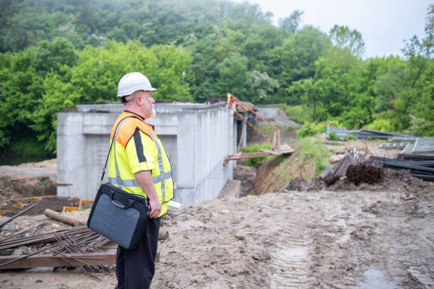 Engineer with a white helmet, white beard, reflective vest, and laptop bag at the bridge construction site early in the morning.