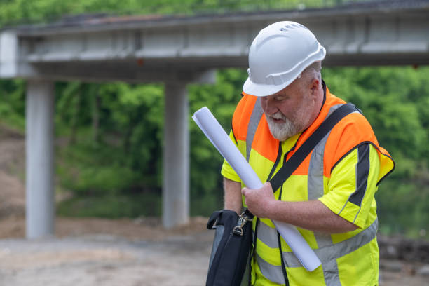 istock Engineer with a white helmet, white beard, reflective vest, and laptop bag at the bridge construction site early in the morning. 2159971918