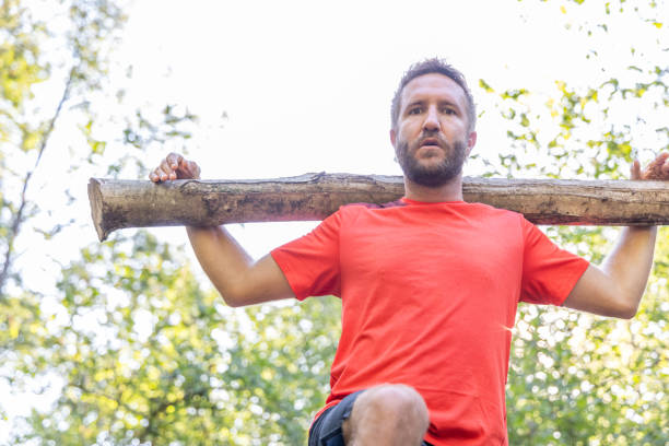 A man in a red shirt and black shorts walks through a forest carrying a log on his shoulder, demonstrating strength and endurance.