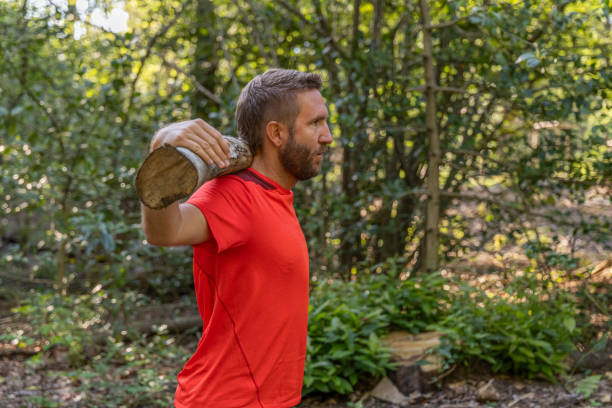 A man in a red shirt performs a squat exercise with a log on his shoulders in a forest setting, showcasing outdoor fitness and strength training.