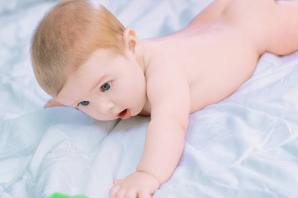 Happy baby crawls on a cozy bed with white sheets, gazing curiously with big blue eyes, surrounded by love in a bright bedroom