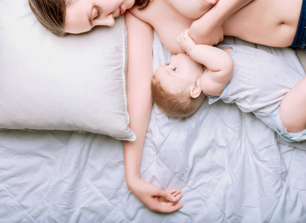 New mom is sleeping soundly in a white bed while breastfeeding her baby, capturing the special bond and tranquility of motherhood