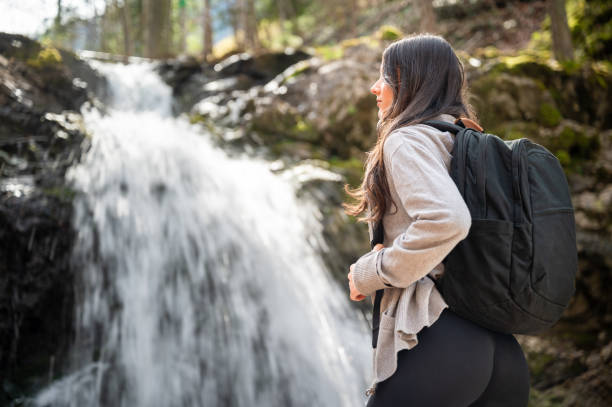 istock Young woman stands on a rocky terrain before a waterfall with threes on the background during a hike 2159995009