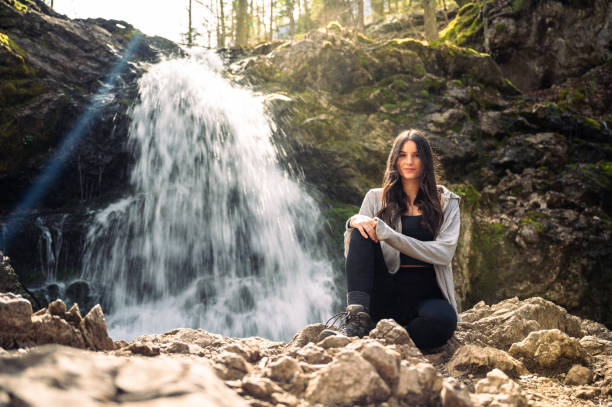 istock Young woman sitson a rocky terrain before a waterfall with threes and moss on the background during a hike 2159995103