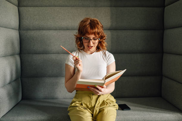 Portrait of young fashionable confident businesswoman sitting at modern workspace with textbook in hands and coming up with creative ideas. Trendy content writer contemplating creative ideas at office