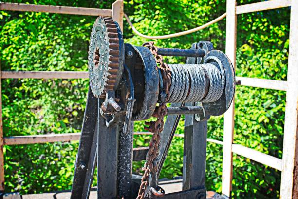 Close-up of a mechanical winch with a steel cable and chain, set against a green leafy background.