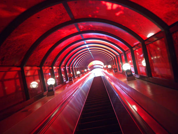 Going up from the dark to the light on Long straight escalator in red glass tunnel with lanterns, perspective view