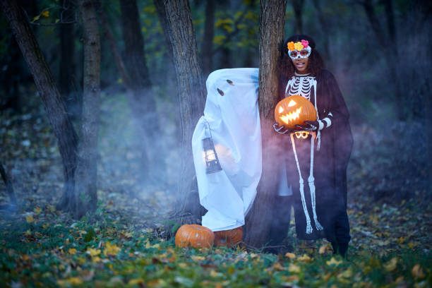 Happy African American girl holding illuminated Jack O' Lantern in the forest while ghost is standing next to her.