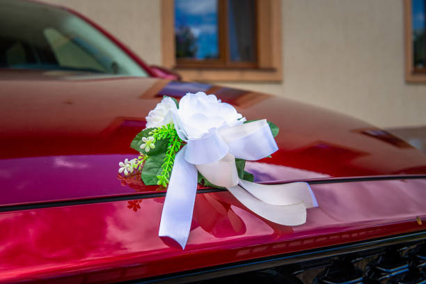 A close-up shot of a white floral arrangement tied with a white ribbon, adorning the hood of a red car. The car is likely the wedding getaway vehicle, waiting outside a reception hall. The flowers are arranged in a bouquet shape and are tied with a large bow, adding a touch of elegance to the occasion.