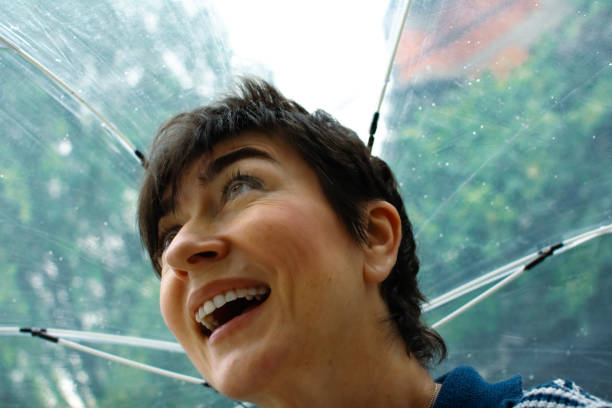 Joyful young girl embracing the rain with pure happiness. She stands outdoors in lush, green nature, arms outstretched, tilting her head back with her tongue out to catch raindrops.  The blurred greenery in the background enhances the natural and carefree atmosphere, making this a heartwarming and lively depiction of childhood joy and connection with nature.