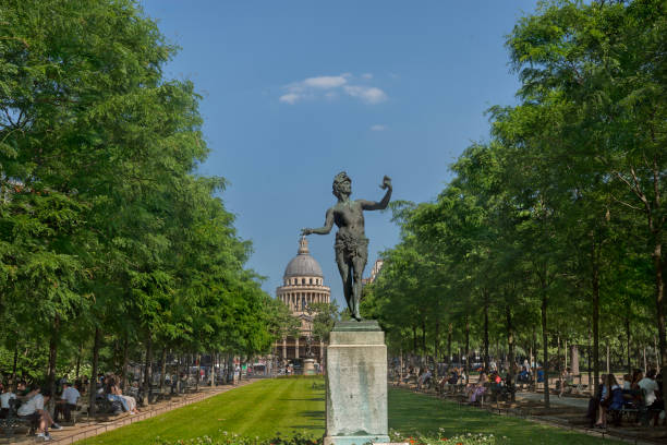 The bronze statue in the Jardin du Luxembourg entitled «LActeur Grec» by Arthur Bourgeois, seen in front of the Panthéon. Bourgeois created the sculpture in 1868 which portrays a Greek theater actor reading the manuscript he holds in his left hand. He wears a mask raised on his forehead and a sheepskin surrounds his midriff.