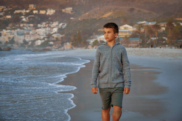 A teenage boy walks alone on the beach during sunset. The boy's expression is contemplative, reflecting the peaceful and introspective atmosphere of the moment. The serene setting, with the calming sounds of the ocean and the warm colors of the sunset, captures a quiet and reflective slice of time.
