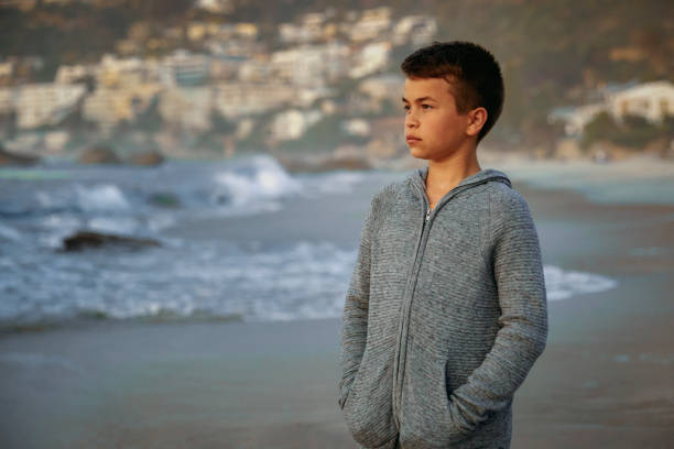 A teenage boy walks alone on the beach during sunset. The boy's expression is contemplative, reflecting the peaceful and introspective atmosphere of the moment. The serene setting, with the calming sounds of the ocean and the warm colors of the sunset, captures a quiet and reflective slice of time.