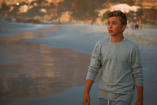 A teenage boy walks alone on the beach during sunset. The boy's expression is contemplative, reflecting the peaceful and introspective atmosphere of the moment. The serene setting, with the calming sounds of the ocean and the warm colors of the sunset, captures a quiet and reflective slice of time.