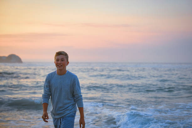 A teenage boy walks alone on the beach during sunset. The serene setting, with the calming sounds of the ocean and the warm colors of the sunset, captures a quiet and reflective slice of time.