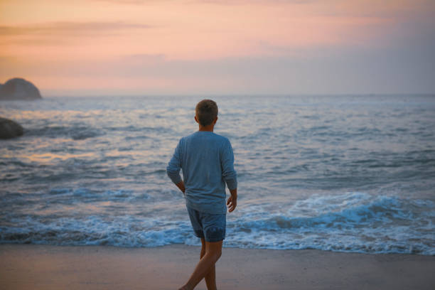 A teenage boy walks alone on the beach during sunset, the sky painted with hues of orange, pink, and purple. His silhouette is softly outlined against the glowing horizon as he strolls along the shoreline, the gentle waves lapping at his feet.