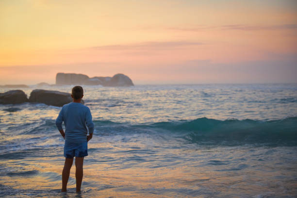 A teenage boy stands alone on the beach during sunset, the sky painted with hues of orange, pink, and purple. His silhouette is softly outlined against the glowing horizon as he stands along the shoreline, the gentle waves lapping at his feet.