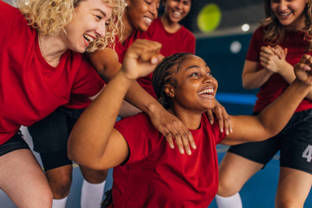 Happy female athletes in matching red shirts embracing and smiling as they celebrate a victory in a sports facility, capturing the essence of camaraderie and teamwork.