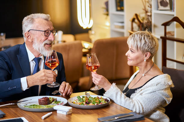 Stylish senior couple taking meal in restaurant, using electronic cigarette. IQOS Stylish, elegant senior couple look at each other and chatting, close relationship. Grey-haired man in tuxedo raise a glass. Use e-cigarette, vape. Modern technologies iqos e cig stock pictures, royalty-free photos & images