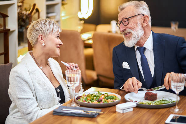 Senior modern couple taking a meal in restaurant using IQOS, electronic cigarette Elderly couple have dinner in rich restaurant while using IQOS, e-cigarette. Handsome man in tuxedo smile, look at woman and speak. iqos e cig stock pictures, royalty-free photos & images