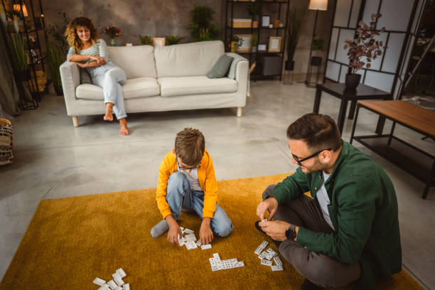 istock Dad and son play dominoes together on the floor while mom sit on sofa 2160089201