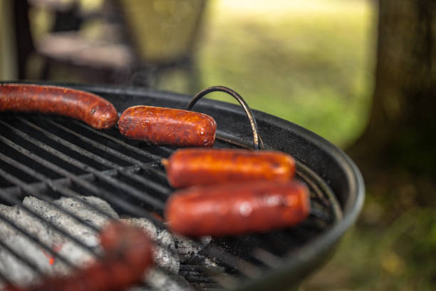 A Bratwurst or hot dog is being displayed on tongs cooked on a Flaming grill, with smoke and flames rising from the grill. A common outdoor activity during the summer months